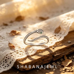 Könnte beinhalten: Ein silberfarbener Schlangenring mit einem strukturierten Schuppenmuster auf der Ringschiene, einem Kopf mit kleinen klaren Steinen und einem einzelnen grünen Stein als Auge. Der Text SHABANAJEWL steht unten im Bild.