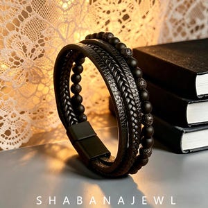 Puede incluir: Una pulsera de múltiples hilos con cordones de cuero trenzado negro y cuentas de piedra negra mate, incluyendo cuentas de roca volcánica texturizada. La pulsera se asegura con un cierre magnético negro rectangular y muestra el texto SHABANAJEWL.