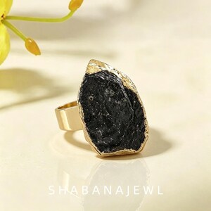 May include: A gold-toned ring featuring a large, irregular, raw black stone centerpiece with a textured surface. The band is smooth and metallic, and the text SHABANAJEWL is printed in white at the bottom of the frame.