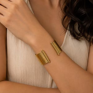 Peut inclure: Un bracelet manchette doré composé d'une série de fines tiges métalliques parallèles disposées en un design incurvé et ouvert. La structure se compose de multiples segments verticaux semblables à des fils reliés aux extrémités.