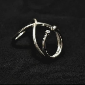 Puede incluir: Un anillo plateado con un diseño único. Presenta una banda curva que envuelve un anillo más pequeño, adornado con dos pequeños adornos redondos. El anillo se encuentra sobre un fondo oscuro, resaltando su brillo metálico.
