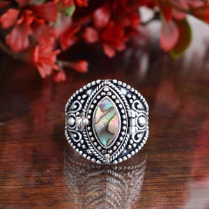 Op de afbeelding: Zilverkleurige ring met een ovale, iriserende abalone schelp middensteen. De ring heeft ingewikkelde details rond de steen en band, met een vintage uitstraling. De ring is een sieraad.