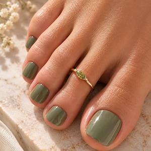 May include: A thin gold toe ring featuring a small, marquise-cut olive green gemstone set in the centre. The ring is worn on the second toe, which is painted with a matching olive green nail polish, consistent with the other toes.
