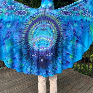 May include: A vibrant blue tie-dye hooded cloak with a circular design in the center, featuring shades of purple and green. The cloak's wingspan is spread out, showcasing the intricate patterns and colors. The cloak is worn over tan pants and gray shoes.