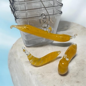 May include: Three yellow glass art pieces, resembling slugs, are displayed on a stone surface. One is suspended from a clear plastic container with a metal handle. The glass pieces have a translucent, textured appearance.