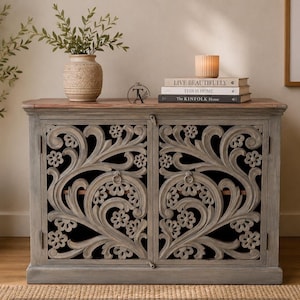 Op de afbeelding: Een rechthoekige houten kast met een verweerde grijze afwerking, voorzien van twee deuren met gedetailleerde bloem- en krulmotieven. De kast heeft een bovenblad van natuurlijk hout en kleine ronde metalen ringgrepen op de deuren.