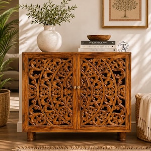 Peut inclure: Un meuble rectangulaire en bois doté de deux portes avec des motifs floraux circulaires sculptés. Le meuble repose sur de petits pieds et possède deux petits boutons ronds. Un vase blanc moucheté et trois livres sont posés dessus.
