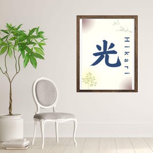May include: A framed art print featuring the Japanese Kanji character for "Hikari" (light) in navy blue. The artwork has a cream background with subtle floral accents and a dark brown wooden frame. The print is displayed in a room with a white chair and a potted plant.