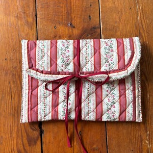 May include: A rectangular quilted fabric pouch featuring a pattern of vertical red stripes alternating with cream-colored bands decorated with small green leaves and tiny red flowers. The pouch has a fold-over flap secured by a thin dark red ribbon tie.