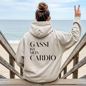 Könnte beinhalten: Beige Kapuzenpullover mit dem Text "GASSI IST MEIN CARDIO" in Schwarz. Die Person, die den Kapuzenpullover trägt, steht auf Holzstufen, mit dem Meer und dem Strand im Hintergrund.