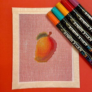 May include: A needlepoint canvas featuring a colorful, stitched mango design. The canvas is surrounded by a beige border and set against a red background. Six acrylic markers in various colors are positioned diagonally across the top right corner.