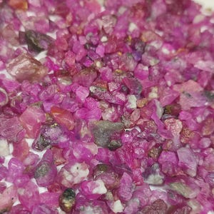 May include: A close-up view of a pile of raw ruby gemstones. The gemstones are a vibrant pink and red hue, with varying shapes and sizes. Some stones have a translucent quality, while others are more opaque. The gemstones are scattered across a white surface.