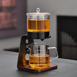 Può includere: Un infusore per tè in vetro con una caraffa in vetro trasparente e un misurino. L'infusore per tè è riempito di liquido ambrato e il misurino ha delle tacche di volume. L'infusore poggia su una base di legno e un telaio in metallo nero.