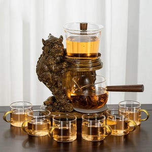 May include: A tea set featuring a decorative, bronze-colored stand with a mythical creature design. The set includes a glass teapot, a serving pot with a wooden handle, a lidded infuser, and several small glass teacups with gold-colored handles, all filled with tea.