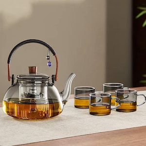 May include: A clear glass teapot with a black handle and a wooden lid, filled with tea. The teapot is accompanied by four small glass teacups, all containing tea. A decorative blue and black charm hangs from the handle.