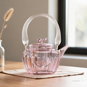 May include: A translucent pink glass teapot featuring a ribbed body, a clear glass handle with a twisted spiral texture, and a lid topped with a small pink glass flower-shaped knob. The teapot includes a matching internal glass infuser basket.