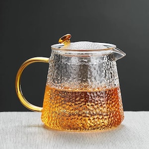 Könnte beinhalten: Eine klare Glas-Teekanne mit strukturierter Oberfläche, einem goldenen Griff und Deckel. Die Teekanne ist mit einer goldbraunen Flüssigkeit gefüllt, wahrscheinlich Tee. Das Design ist elegant und zum Servieren von Tee geeignet.
