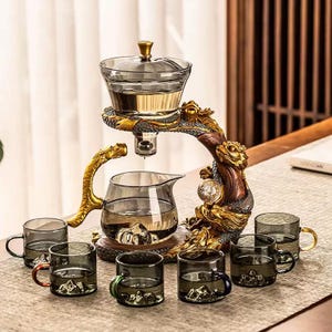 May include: A tea set featuring a glass teapot and cups, with a decorative golden dragon-shaped stand. The set includes a teapot with a lid, a serving pot, and six small cups, all in a smoky gray color. The dragon stand adds an ornate touch.