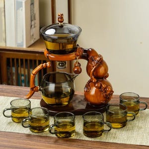 Puede incluir: Un juego de té de madera con una tetera de vidrio, un infusor de vidrio y seis tazas de té pequeñas de vidrio. El juego tiene un acabado de madera oscura y está dispuesto sobre una bandeja de madera. El juego está diseñado para preparar y servir té.