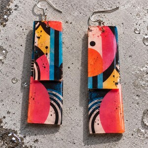 Puede incluir: Un par de pendientes rectangulares colgantes con un diseño abstracto vibrante de formas geométricas superpuestas en tonos rosa intenso, naranja brillante, amarillo y azul claro, acentuados con líneas y motas negras, con ganchos plateados.