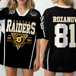Op de afbeelding: Zwart en wit Boston Raiders-shirt met de naam "ROZANOV" en het nummer "81" op de rug. De voorkant heeft de teamnaam en het logo met de tekst "EST. 1924". Het shirt heeft een V-hals en witte strepen.