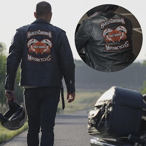 May include: Black leather motorcycle jacket with Harley-Davidson logo patches on the back. The patches feature the Harley-Davidson name and the word "Motorcycles" in white and orange lettering. A motorcycle helmet is held in the hand.