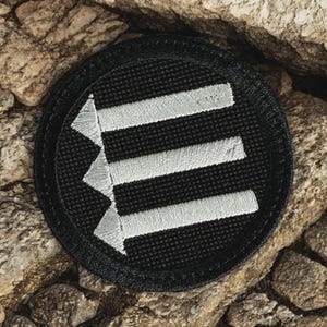 May include: A round black patch with three horizontal white arrows pointing left. The patch is embroidered with a textured design and has a black border. The patch is set against a background of rough, brown rocks.