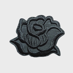 May include: A black embroidered rose patch. The patch features a detailed design of a rose with multiple petals and leaves. The patch is made of a dark fabric and has a textured appearance. The rose is a classic floral design.