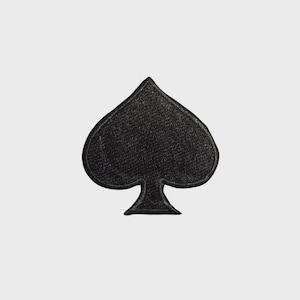 May include: A black spade patch, a playing card symbol, is centered against a white background. The patch has a textured appearance and a classic spade shape, with a rounded top and a stem at the bottom.