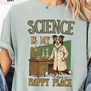 Könnte beinhalten: Ein helles salbeigrünes T-Shirt mit einer Grafik eines Hundes im weißen Laborkittel und mit Schutzbrille, der an einem Holztisch mit Globus, Büchern und Bechergläsern steht. Der Text lautet SCIENCE IS MY HAPPY PLACE in grüner und gelber Schrift.