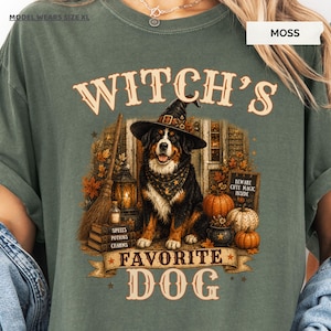 May include: A moss green t-shirt featuring a graphic of a Bernese Mountain Dog wearing a witch hat and bandana. The design includes a broom, stacked books labeled Spells, Potions, Charms, pumpkins, and a sign reading Beware Cute Magic Inside.