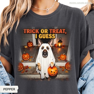 Può includere: Una t-shirt grigio antracite con la grafica di un pastore tedesco che indossa un costume da fantasma bianco. Il cane tiene in mano un secchiello a forma di zucca arancione su un portico con zucche e una lanterna. Il testo recita: TRICK OR TREAT, I GUESS.