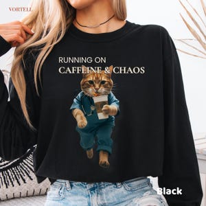 Peut inclure: Sweat-shirt noir avec le texte "RUNNING ON CAFFEINE & CHAOS". Le graphisme représente un chat portant une blouse et tenant une tasse de café. Le chat court. Le mot "Black" est imprimé en bas à droite.