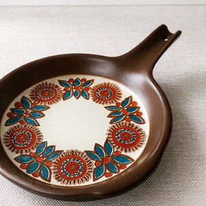 May include: Brown ceramic serving dish with a handle. The dish features a white interior with a floral design in red, blue, and brown. The handle has a hole for hanging.