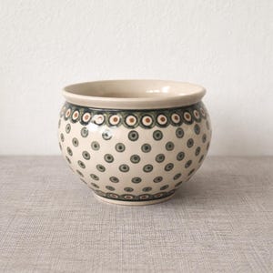 Może przedstawiać: Okrągła ceramiczna miska z kremową podstawą i dekoracyjnym wzorem zielonych i brązowych kropek. Obrzeże ma obramowanie z naprzemiennych zielonych, brązowych i białych kół. Miska ma średnicę około 15 cm.