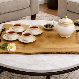 May include: A tea set on a wooden serving tray. The set includes a white teapot with gold detailing, four teacups and saucers, and a small bowl of tea leaves. A daisy and a flower stem are on the tray.