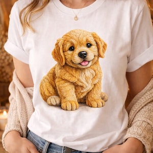May include: White t-shirt featuring a golden retriever puppy illustration. The puppy is sitting, with a happy expression and its tongue sticking out. The shirt is worn by a person wearing blue jeans.