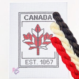 May include: A white needlepoint canvas featuring a black outlined rectangular border, the word CANADA, a red maple leaf graphic, and the text EST. 1867. Three twisted embroidery floss skeins in dark brown, cream, and red are placed on the right side.