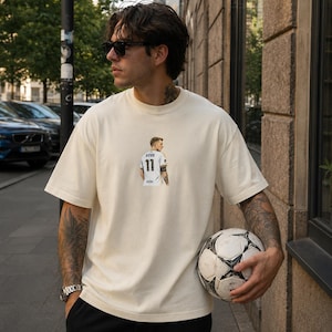 Peut inclure: Un t-shirt à manches courtes blanc cassé avec un petit motif sur la poitrine représentant un joueur de football portant un maillot blanc avec le nom REUS et le numéro 11 imprimés au dos. Le t-shirt a un col rond et une coupe décontractée.