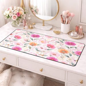 May include: A rectangular desk mat featuring a floral pattern with pink and orange flowers, green leaves, and a white background. The mat is bordered with a thin black edge. The mat is placed on a white desk.