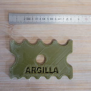 Peut inclure: Outil en argile vert olive avec le mot "ARGILLA" imprimé. L'outil a un bord ondulé et un trou central. Une règle métallique est placée au-dessus de l'outil, indiquant les mesures en centimètres.