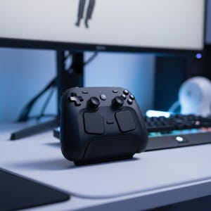 Könnte beinhalten: Ein schwarzer Gaming-Controller mit mehreren Tasten und zwei quadratischen Pads. Der Controller liegt auf einer weißen Oberfläche, mit einem Computermonitor und einer Tastatur im Hintergrund. Der Controller hat ein modernes Design.