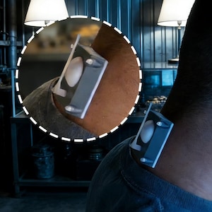 May include: A small, rectangular device with a white oval shape in the centre is attached to a person's arm and shoulder. The device is grey and has clear, rounded supports. The background is a dimly lit room.