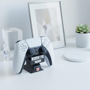 Op de afbeelding: Een wit en zwarte PlayStation 5-controller op een zwarte standaard. De standaard toont de tekst "RESIDENT EVIL requiem" en een rood-wit paraplu-logo. Een ingelijste foto en een kleine potplant staan op de achtergrond.