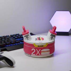 May include: A pink and white burger-shaped device with two pink mouth-shaped attachments. The device has the text "2X BURGER" and a burger graphic. A keyboard and game controller are in the background.