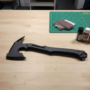 Pode incluir: Um machado tático tomahawk preto com um cabo texturizado e uma lâmina afiada. O machado tem um espinho no lado oposto da lâmina. Está sobre uma superfície de madeira. Lixas e frascos de tinta também são visíveis.
