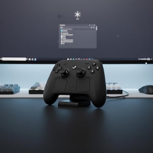 Könnte beinhalten: Ein schwarzer Gaming-Controller mit Steam-Logo, auf einem schwarzen Ständer. Der Controller hat zwei Joysticks, Tasten und ein Steuerkreuz. Ein Computermonitor und eine Tastatur befinden sich im Hintergrund.