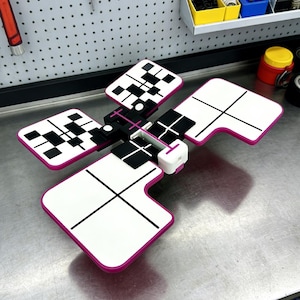May include: A white and pink geometric tool with black accents. The tool has four rectangular panels with black crosshairs and checkered patterns. The tool is on a metal surface.