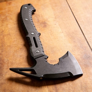 Puede incluir: Un hacha táctica tomahawk gris oscuro con un mango curvo y una hoja afilada. El hacha presenta un agarre texturizado, múltiples detalles de pernos y un extremo puntiagudo opuesto a la hoja. El hacha se muestra sobre una superficie de madera.