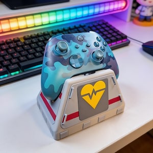 May include: A teal and blue camouflage Xbox controller rests on a gray charging stand with a yellow heart and heartbeat graphic. The stand has red stripes and is on a white surface. A keyboard and colorful LED lights are in the background.